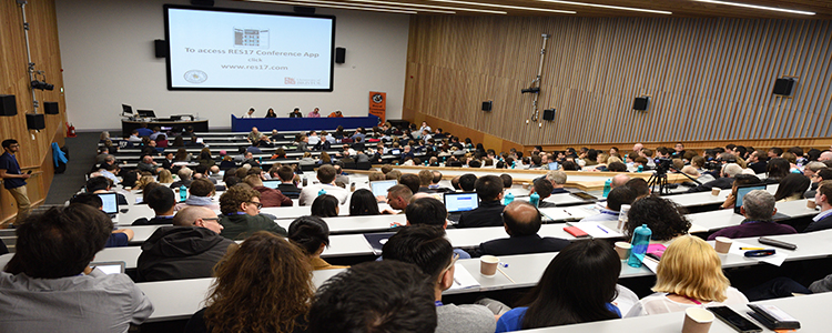 A lecture theatre with delegates, watching a presentation on projector and panel of 5 speakers.